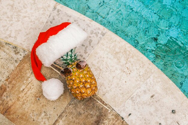 Foto grappige ananas met zonnebril en kerstman hoed tegen zwembad en tropische planten in zonnige wea