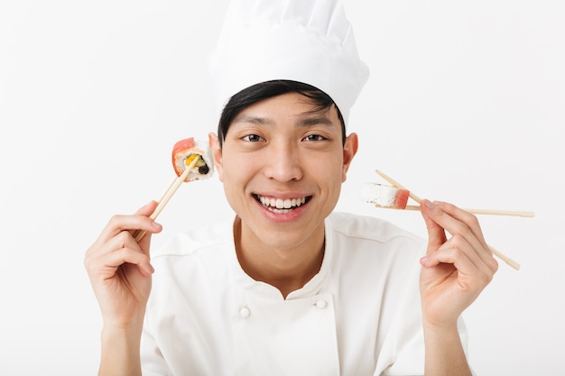 Grappige aziatische belangrijkste man in witte kok uniforme eten sushi set met stokjes geïsoleerd over witte muur