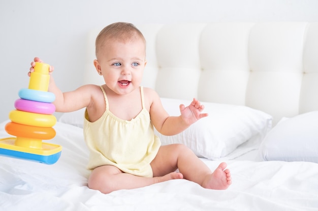 Grappige babymeisje in gele bodysuit spelen met plastic speelgoed kinderen piramide op bed