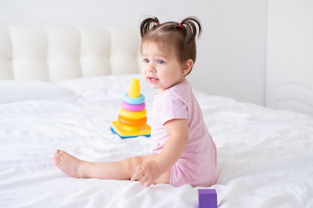 grappige babymeisje in roze bodysuit spelen met plastic speelgoed kinderen piramide op bed