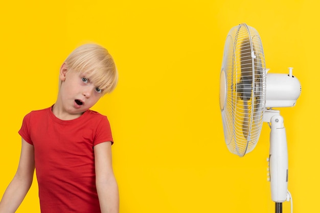 Grappige blonde jongen vormt gezichten in de buurt van de ventilator Gele achtergrond Hete zomer