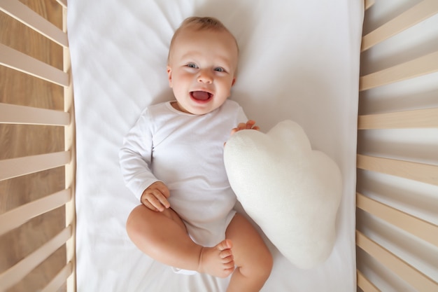 Grappige blonde schattige kleine blauwogige baby in witte Romper liggen en lachen in wit kinderbed, knuffels wolk kussen, kopie ruimte, horizontaal, bovenaanzicht