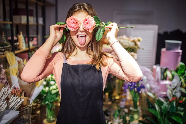 Grappige en positieve jonge vrouwelijke bloemist bedekken ogen met rozen en tonen tong. Ze heeft lol. Planten en bloemen staan achter in vazen.