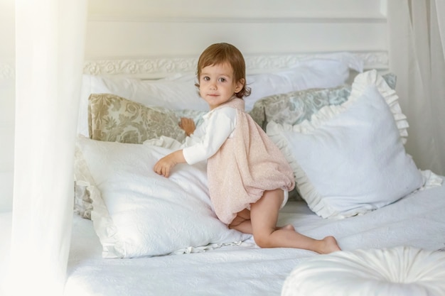 Grappige en schattige brunette lachend meisje spelen springen op bed in lichte slaapkamer. Wit interieur met groot bed. Jeugd, kleuterschool, jeugd, ontspannen concept