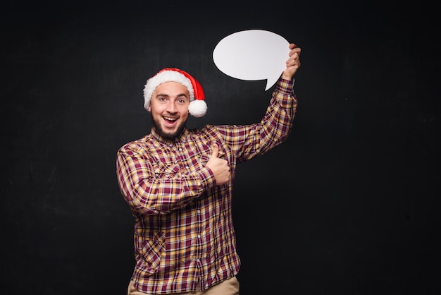 Grappige ernstige man in rode kerst-kerstmuts houdt leeg wit karton als blanco of bespotten met kopie ruimte voor tekst. Zwarte achtergrond