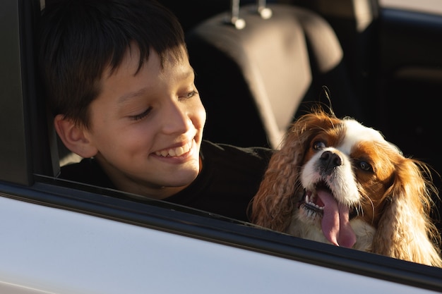Grappige foto van een tienerjongen die lacht en uit het autoraam kijkt met zijn hond cavalier king charles spaniel, op het punt om op reis te gaan