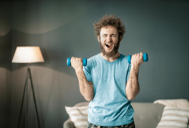 Grappige gekrulde man geniet ervan zijn biceps te pompen door twee speciale halters grappige man in blauwe tshirt heeft spor...