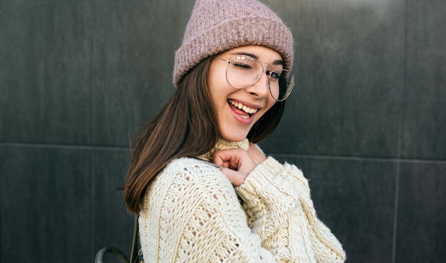 Grappige glimlachende jonge vrouw knippert met een oog en kijkt naar de camera met een gebreide trui, een transparante bril en een hoed die zich voordeed op de straat van de stad