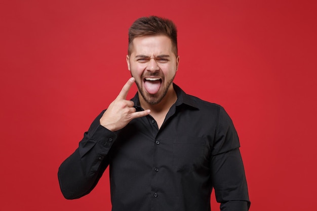 Grappige jonge bebaarde man 20s in klassieke zwarte shirt poseren geïsoleerd op rode achtergrond. Mensen levensstijl concept. Bespotten kopie ruimte. Beeltenis van heavy metal rock teken, hoorns omhoog gebaar, tong tonen.