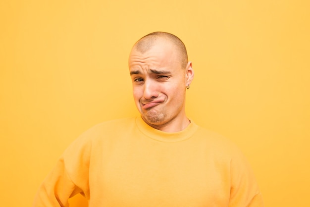 Grappige jonge man in een geel sweatshirt maakt een grappig gezicht en kijkt naar de camera op een geel