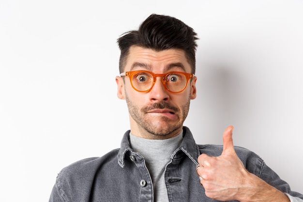 Foto grappige jonge man in glazen duimen opdagen, lip bijten en besluiteloos kijken, staande op een witte achtergrond.