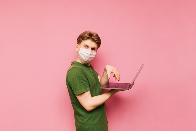 Grappige jonge man met een beschermend medisch masker geïsoleerd op een roze achtergrond met een laptop in zijn hand kijken naar de camera met een kwaad gezicht en typen op het toetsenbord quarantaine concept