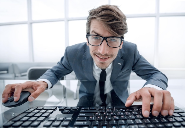 Grappige jonge man met een gek gezicht zittend aan de tafel met handen op muis en computertoetsenbord