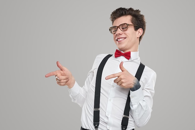 Grappige jonge mannelijke student in nerdy outfit en bril die opzij wijst en lacht tegen een grijze achtergrond