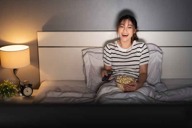 Grappige jonge vrouw die tv kijkt en 's nachts op een bed lacht