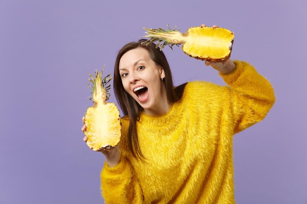 Grappige jonge vrouw in bonttrui die mond open houdt met helften van vers rijp ananasfruit dat op violette pastelachtergrond wordt geïsoleerd. Mensen levendige levensstijl ontspannen vakantie concept. Bespotten kopie ruimte.