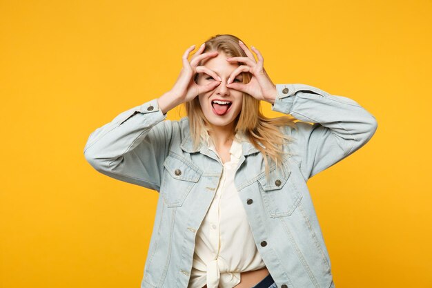 Grappige jonge vrouw in denim vrijetijdskleding houdt handen in de buurt van ogen, imiteert een bril of verrekijker, toont tong geïsoleerd op geeloranje muurachtergrond. Mensen levensstijl concept. Bespotten kopie ruimte.