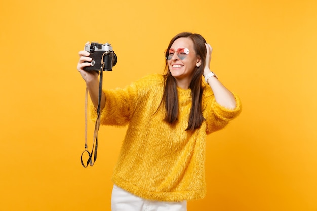 Grappige jonge vrouw in hart bril doen selfie geschoten op retro vintage fotocamera, hand op het hoofd geïsoleerd op gele achtergrond te zetten. Mensen oprechte emoties, levensstijl. Reclame gebied.