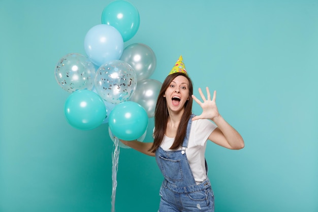 Grappige jonge vrouw in verjaardagshoed die mond open houdt met palm en viert, met kleurrijke luchtballonnen geïsoleerd op blauwe turkooizen achtergrond. Vakantie verjaardagsfeestje, mensen emoties concept.