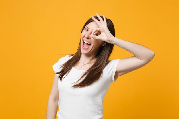 Grappige jonge vrouw in witte casual kleding hand in hand in de buurt van ogen, imitatie van een bril of verrekijker geïsoleerd op fel geel oranje achtergrond in de studio. mensen levensstijl concept. bespotten kopie ruimte.