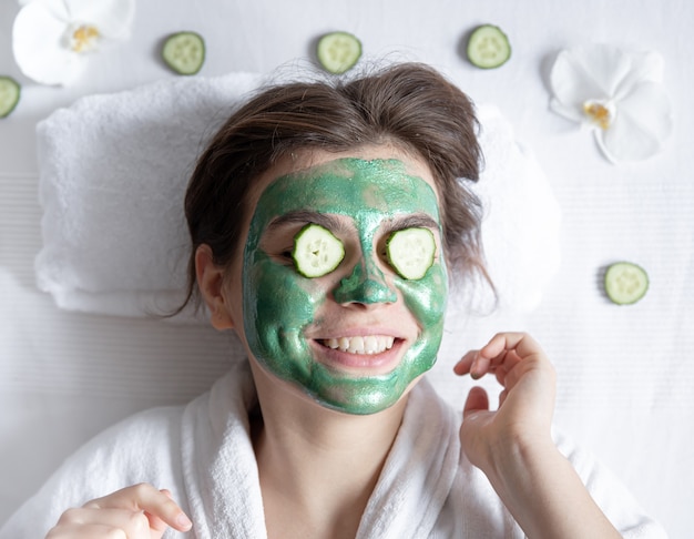 Grappige jonge vrouw met een cosmetisch masker op haar gezicht en komkommers op haar ogen.