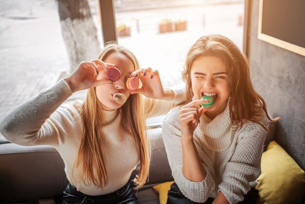 Grappige jonge vrouwen bedekken ogen met macarons. Ze poseren op camera. Brunette bijt stuk macarone. Zij hebben lol.