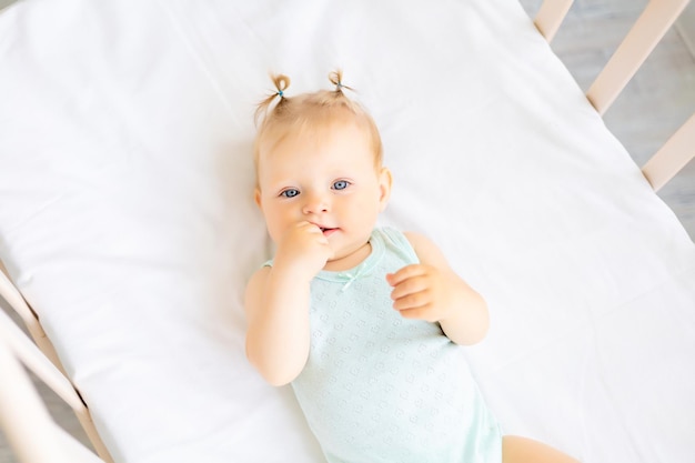 Grappige kleine baby die in een wieg staat in een witte kinderkamer met een raam een klein meisje in een katoenen bodysuit glimlachende blauwogige baby