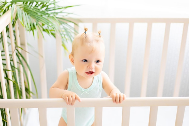 Grappige kleine baby die in een wieg staat in een witte kinderkamer met een raam Een klein meisje in een katoenen bodysuit Glimlachende blauwogige baby