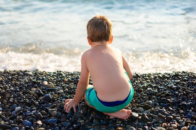 Grappige kleine babyjongen in korte broek op het strand Het kind zit met zijn rug tegen de achtergrond van de zee Gelukkig klein kind op zomervakantie