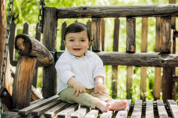 Grappige kleine jongen zit in de tuin op een houten schommelbank en lacht om de camera