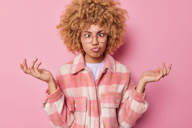 Foto grappige krullende vrouw maakt grimas kruist ogen en pruilt lippen spreidt handpalmen voelt aarzelend draagt transparante bril en geruite jas poses tegen roze achtergrond menselijke gezichtsuitdrukkingen