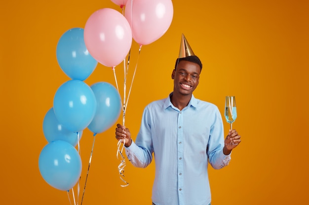 Grappige man in pet met een glas drank, gele achtergrond. Glimlachende mannelijke persoon kreeg een verrassing, evenement of verjaardagsfeestje, ballonnendecoratie