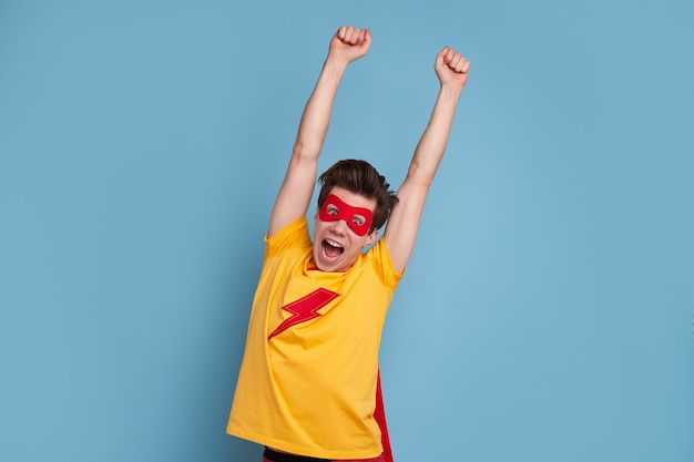 Grappige man in superheld masker schreeuwen en armen opheffen tijdens het vieren van de overwinning tegen blauwe achtergrond