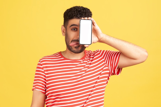 Grappige man met baard in rood gestreepte tshirt plezier en gek rond tonen tong die betrekking hebben op gezicht met witte display smartphone Indoor studio opname geïsoleerd op gele achtergrond