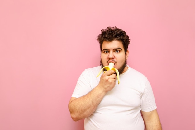 grappige man met overgewicht met krullend haar die banaan eet op een roze achtergrond en in de camera kijkt