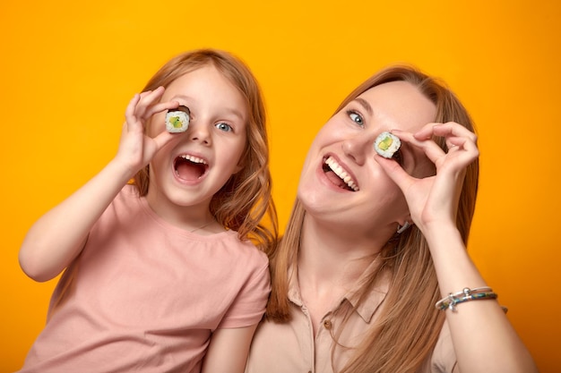 Grappige moeder met dochter die sushibroodjes voor ogen op gele achtergrond houdt