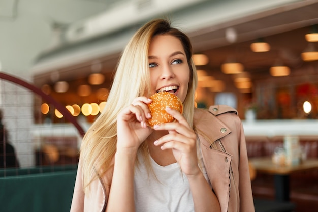 Grappige mooie jonge vrouw in een roze jasje een hamburger eten in een café