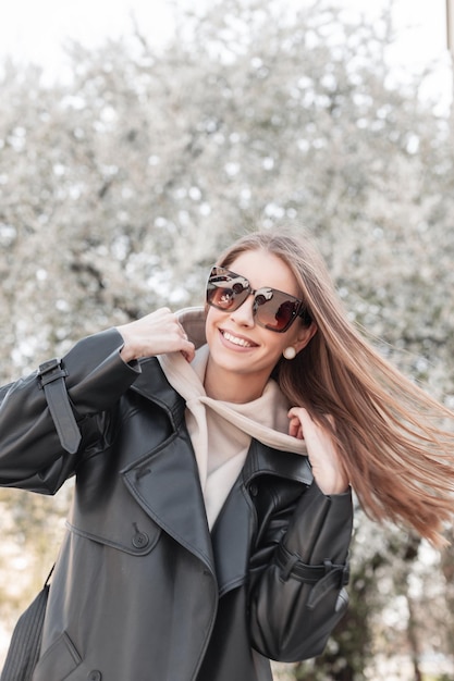 Grappige mooie modieuze gelukkig hipster vrouw model met een glimlach met modieuze zonnebril in een stijlvolle leren lange jas met een hoodie wandelen in de stad in de buurt van een bloeiende boom