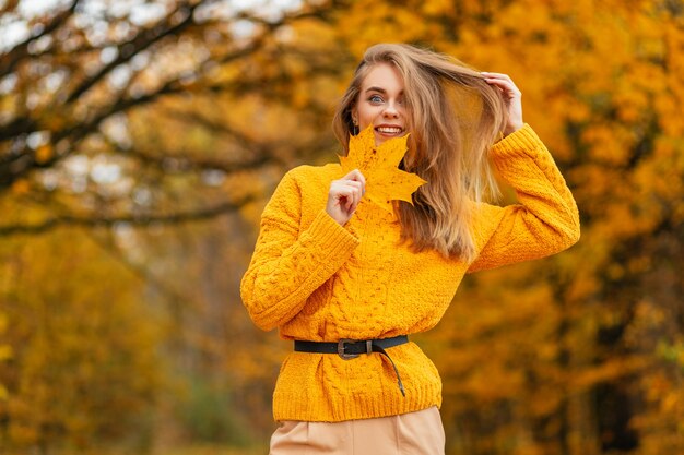 Grappige mooie vrouw met een glimlach in een modieuze gebreide gele trui met een herfstblad in de natuur met gekleurd herfstblad op een herfstdag