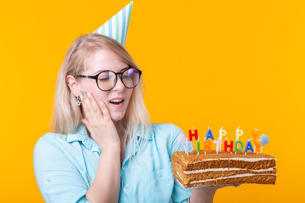 Grappige positieve jonge vrouw houdt in haar handen een zelfgemaakte cake met de inscriptie gelukkige verjaardag poseren op een gele muur. Concept van vakanties en jubilea.