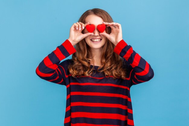 Grappige positieve vrouw dragen gestreepte casual stijl trui die ogen bedekt met kleine rode harten liefde symbool glimlachend tandachtig verliefd Indoor studio shot geïsoleerd op blauwe achtergrond