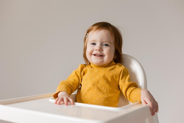 grappige roodharige baby met blauwe ogen in een gele bodysuit zittend in een witte kinderstoel