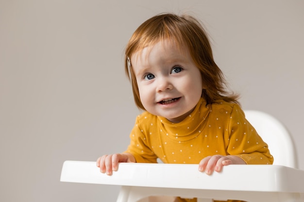 grappige roodharige baby met blauwe ogen in een gele bodysuit zittend in een witte kinderstoel