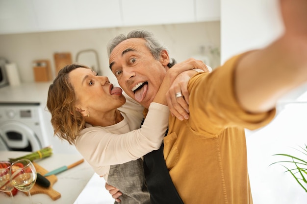 Grappige senior paar maken selfie knuffelen en grimassen staan in keuken interieur en plezier hebben