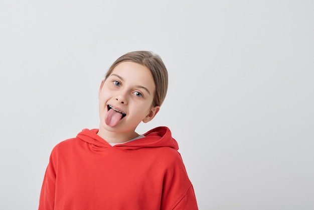 Grappige tiener in rood sweatshirt die tong toont terwijl het grimassen voor camera tegen witte achtergrond afzonderlijk trekt