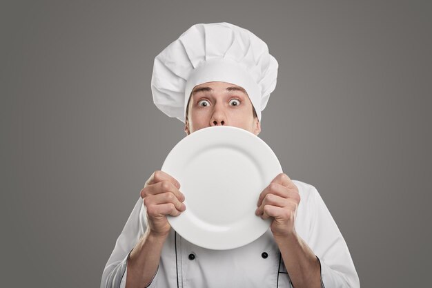 Grappige verbaasde jonge mannelijke kok in uniform van de chef-kok en hoed met lege witte plaat en kijkend naar camera tegen een grijze achtergrond