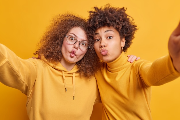 Grappige vrouwen van gemengd ras omarmen en maken grimas, houden de lippen gevouwen in een kus, maken vissenlippen dwaas gekleed in gele kleding, maken selfie vriendelijke relaties. Vriendschap