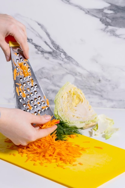 grated carrots on an iron grater lie on a yellow board next to cabbage and greens