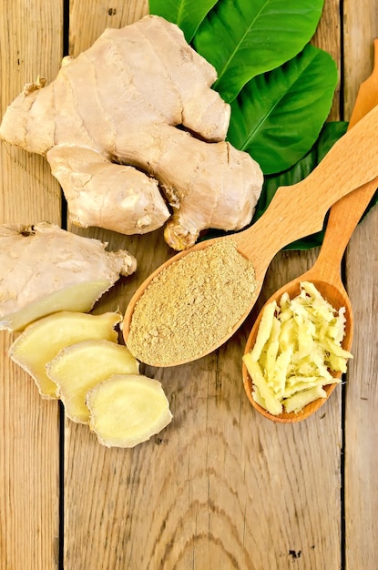 Grated and ground ginger in two wooden spoons, ginger root, green leaves on the background of wooden board