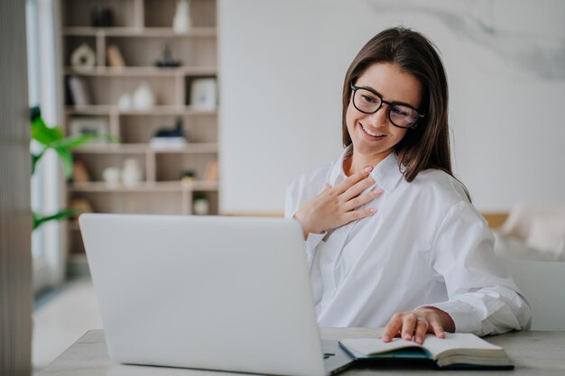 Foto grata giovane imprenditrice con gli occhiali della camicia bianca seduta al tavolo in ufficio che effettua videochiamate